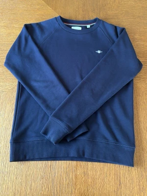 Mörkblå sweatshirt från GANT - Klassisk mörkblå sweatshirt från GANT med rund halsringning och diskret logga broderad på bröstet. Tröjan har ribbade muddar vid ärmslut och nederkant. Perfekt för en avslappnad och stilren look.