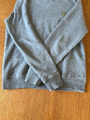 Grå sweatshirt med gul logga. Lyle and scott(xs) - En stilren grå sweatshirt med rund hals och ribbade muddar. På bröstet sitter en liten gul logga som detalj. Perfekt till jeans eller joggers för en avslappnad look. Materialet känns mjukt och bekvämt.