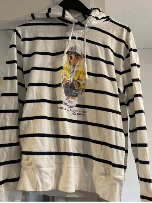 Randig hoodie med Polo Bear-motiv - Vit hoodie med svarta horisontella ränder från Ralph Lauren. På bröstet finns ett stort Polo Bear-tryck där björnen har på sig en gul jacka och keps. Hoodien har huva med dragsko och långa ärmar. Perfekt för en avslappnad och trendig stil.