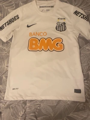Vit Santos Neymar Jr matchtröja - Säljer en vit Santos FC matchtröja med Neymar Jr och nummer 11 på ryggen. Tröjan har tryckta sponsorer som BMG och Netshoes, samt klubbmärke och Nike-logga på bröstet. Tillverkad i Dri-FIT material som andas, perfekt för fotbollsträning eller match.