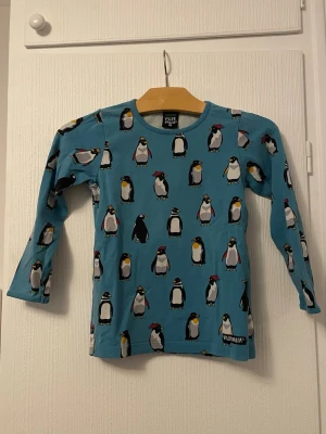 Blå långärmad topp med pingviner - Blå långärmad topp från Viller Valla med ett gulligt pingvinmönster över hela tröjan. Tillverkad i mjuk ekologisk bomull, perfekt för dig som gillar färgglada och lekfulla plagg. Rund halsringning och normal passform.