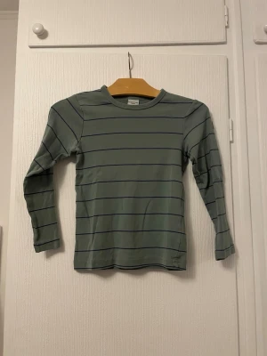 Randig långärmad topp Polarn O. Pyret - Grön långärmad topp från Polarn O. Pyret med tunna mörkblå ränder. Klassisk rund halsringning och mjukt material som känns skönt mot huden. Perfekt för en avslappnad och stilren look.