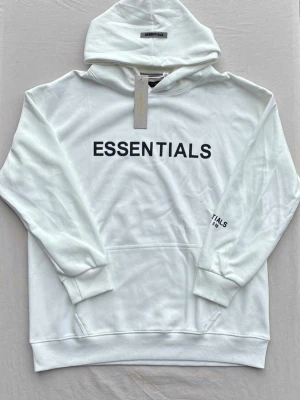 Vit Essentials hoodie med tryck - Säljer en stilren vit hoodie från Essentials med svart tryck på bröstet och liten logga på ärmen. Hoodien har huva, ribbade muddar och magficka. Perfekt för dig som gillar enkel och clean streetwear-look.