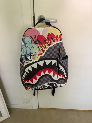 Färgglad ryggsäck med hajmotiv - Cool ryggsäck med rutigt mönster i grått och svart, färgglada graffiti-detaljer och stort hajmotiv framtill. Ryggsäcken har dragkedja, sidofickor och justerbara axelband. Perfekt för dig som vill sticka ut med en unik och trendig väska.