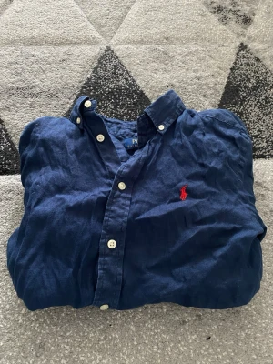 Blå linneskjorta från Polo Ralph Lauren - Snygg blå skjorta i linne från Polo Ralph Lauren med klassisk krage och knappar framtill. Skjortan har den ikoniska röda loggan broderad på bröstet och passar perfekt till en avslappnad men stilren look.