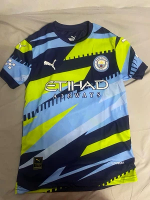 Manchester City matchtröja Puma - Snygg Manchester City matchtröja från Puma med grafiskt mönster i blått, ljusblått och neongult. Tröjan har klubbmärke på bröstet, Etihad Airways-tryck och korta ärmar. Perfekt för fotbollsträning eller att bära på matchdag.