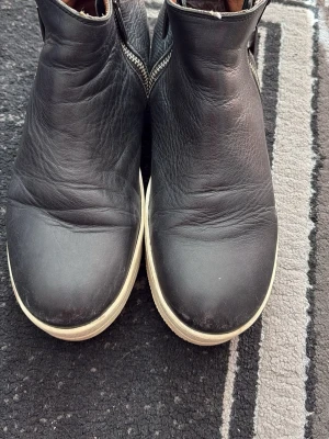 Svarta Chelsea boots i skinn - Säljer ett par klassiska svarta Chelsea boots i skinn med vit sula. Bootsens ovandel är slät och har resår på sidorna för enkel på- och avtagning. Insidan är fodrad i brunt och sulan är något tjockare för en modern look.