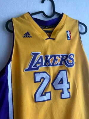 Gul Lakers basketlinne nr 24 - Säljer en gul och lila basketlinne från Adidas med Lakers-tryck och nummer 24 på både fram- och baksidan. På ryggen står det 'Bryant'. Linnet har broderade detaljer och är gjort i ett ventilerande polyestermaterial. Perfekt för basketträning eller som supporterplagg. Storleken är L men mer som M!