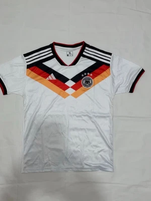 Tysklands fotbollströja retro M - Säljer en retroinspirerad Tyskland fotbollströja i vitt med klassiska svarta, röda och gula detaljer över bröstet. Tröjan har korta ärmar, V-ringning och broderad landslagsemblem med fyra stjärnor. Tillverkad i lätt och ventilerande material.