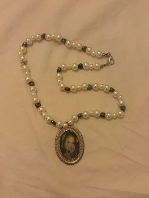Pärlhalsband med vintage medaljong - Unikt halsband med vita pärlor och små silverfärgade detaljer. Medaljongen är oval, silverfärgad och har ett inramat foto i svartvitt. Perfekt för dig som gillar retrostil och personliga accessoarer.