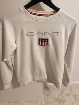 Vit sweatshirt från GANT - Vit sweatshirt från GANT med broderad logga och amerikanskinspirerat märke på bröstet. Tröjan har rund halsringning och ribbade muddar vid ärmslut och nederkant. Klassisk och stilren design med sportig känsla.