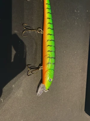 Grön och orange fiskedrag - Säljer ett fiskedrag i plast med grön och orange färg, svarta ränder och silverfärgat öga. Draget har tre trekrokar och en genomskinlig sked framtill. Perfekt för dig som gillar fiske och vill ha ett färgglatt bete i lådan.