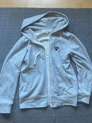 CDG hoodie - Grå zip-hoodie med huva och dragsko, samt fickor på sidorna. På bröstet och ryggen finns en svart hjärtformad detalj med ögon. Hoodien har en avslappnad passform och är tillverkad i mjukt sweatshirtmaterial, perfekt för en chill look. Storlek L sitter ganska litet, hör av er vid frågor!