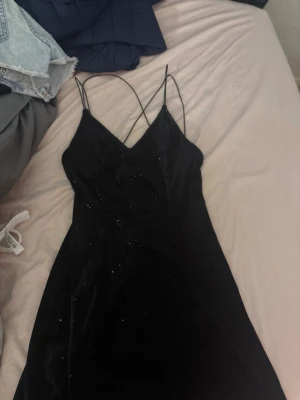 Svart glittrig klänning med tunna band - Snygg svart klänning med glittriga detaljer och tunna axelband som korsas i ryggen. Klänningen har en v-ringad front och är ärmlös, perfekt för dig som vill sticka ut med en elegant look.