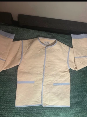 Krämvit quiltad loungewear-jacka - Krämvit quiltad loungewear-jacka från NA-KD med ljusblå kantband och detaljer. Jackan har tryckknappar framtill, två fickor och vida ärmar. Perfekt för mysiga hemmakvällar. Materialet ser ut att vara mjukt och bekvämt.