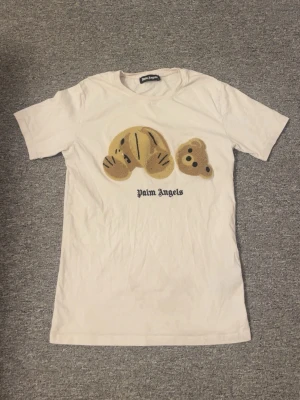 Vit Palm Angels t-shirt med björn - Vit t-shirt från Palm Angels med ett broderat motiv av en nallebjörn på bröstet och texten 'Palm Angels' under. T-shirten har korta ärmar och en klassisk passform. Perfekt för dig som vill ha något trendigt och lekfullt i garderoben.