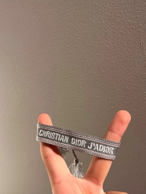 Grått Christian Dior J'Adior armband - Snyggt grått vävt armband från Christian Dior med texten 'J'Adior' broderad i vitt. Armbandet har justerbara band och dekorativa tofsar i ändarna. Perfekt accessoar för att lyfta din outfit med en exklusiv touch.