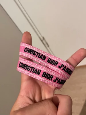 Rosa armband Christian Dior - Säljer ett rosa tygarmband från Christian Dior med texten 'CHRISTIAN DIOR J'ADIOR' i svart. Armbandet har en enkel och trendig design med justerbar knytning. Perfekt accessoar för att lyfta din outfit.