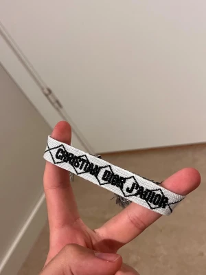 Christian Dior J'Adior tygarmband - Säljer ett vitt tygarmband från Christian Dior med svart broderad text 'J'Adior'. Armbandet har ett grafiskt mönster och justerbara band med fransar i ändarna. Perfekt accessoar för att lyfta din outfit.