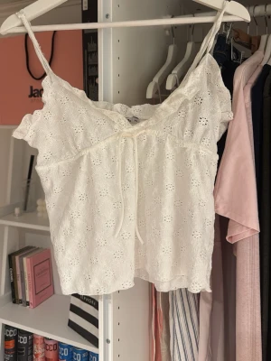 Vit broderad linnetopp med knyt - Söt vit linnetopp med broderade blommor och tunna axelband. Toppen har en liten knytning framtill och volangkant upptill. Perfekt för varma dagar och ger en romantisk vibe till din outfit.