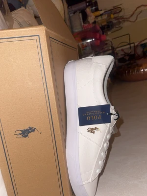 Vita sneakers från Polo Ralph Lauren - Snygga vita sneakers från Polo Ralph Lauren med klassisk låg profil och ikoniska loggan broderad på sidan. Skorna har vita snören och en stilren design i canvasmaterial med vit sula. Perfekta för en clean och fräsch look.