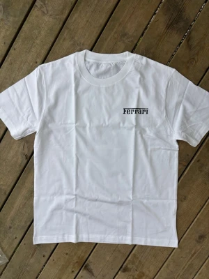 Charles leclerc f1 Vit Ferrari t-shirt med tryck - Snygg vit t-shirt med Ferrari-logga på bröstet och ett stort färgglatt tryck på ryggen som visar en person i röd overall vid en hamn. Klassisk passform och rund halsringning. Perfekt för dig som gillar streetwear och coola prints.