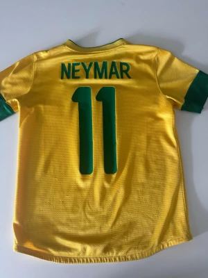 Gul Neymar Brasilien fotbollströja - Gul Brasilien fotbollströja med gröna detaljer, Neymar och nummer 11 på ryggen. Tillverkad i lätt och ventilerande material, perfekt för fotbollsträning eller match. Autentisk Nike-tröja med märkning på nederkanten.