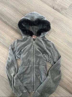 Grå Juicy Couture hoodie med fuskpäls - Säljer en grå hoodie från Juicy Couture i mjuk velour med dragkedja framtill och två fickor. Huvan är kantad med svart fuskpäls för extra stilpoäng. Perfekt för dig som gillar en trendig och bekväm look. Det står att det är xl men den är mindre i storleken. Pälsen är påsydd