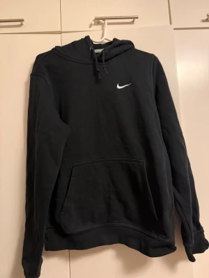 Svart Nike hoodie med logga - Svart hoodie från Nike med vit broderad swoosh-logga på bröstet. Klassisk känguruficka framtill och justerbar huva med dragsko. Mjuk insida och ribbade muddar vid ärmslut och nederkant. Perfekt för en avslappnad streetwear-look.
