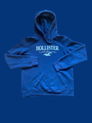 Hollister hoodie (S) - Snygg blå hoodie från Hollister med vit logga och texten 'California' på bröstet. Klassisk känguruficka framtill, ribbade muddar och huva med dragsko. Tillverkad i mjukt bomullsmaterial som är skönt att ha på sig. Perfekt för en avslappnad och cool stil. Kontakta vid intresse!!! OBSERVERA ATT BILDEN ÄR TAGEN I SOLLJUS, VILKET KAN PÅVERKA FÄRGÅTERGIVNINGEN.