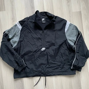 Svart och grå vindjacka från Nike - Snygg svart vindjacka från Nike med gråa paneler på ärmarna och vita detaljer. Jackan har halv dragkedja framtill, hög krage och justerbar dragsko i nederkant. Perfekt för en sportig och avslappnad stil. Nike-logga broderad på bröstet.