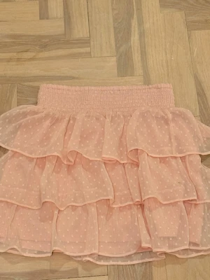 Rosa volangkjol med prickar  - Söt kort rosa kjol med volanger och små prickar. Kjolen har bred resår i midjan och är gjord i ett lätt, transparent material som ger en luftig känsla. Perfekt för dig som gillar en romantisk och lekfull stil.