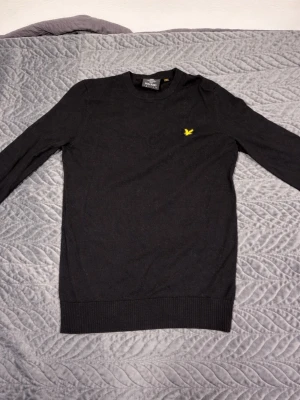 Svart tröja från Lyle & Scott - Svart långärmad tröja från Lyle & Scott med rund halsringning och klassisk gul logga broderad på bröstet. Tröjan har ribbade muddar vid ärmslut och nederkant. Perfekt för en stilren och enkel look.