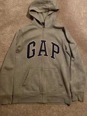Grå zip hoodie från GAP - Säljer en grå hoodie från GAP med dragkedja och stor logga framtill. Hoodien har huva, kängurufickor och mjukt, fleeceliknande innerfoder. Perfekt för chill dagar eller när du vill ha en enkel och snygg look. Storlek: 13år 