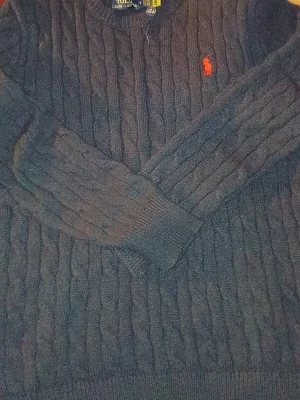 Mörkblå kabelstickad tröja Polo Ralph Lauren - Kabelstickad mörkblå tröja från Polo Ralph Lauren med klassisk röd logga broderad på bröstet. Tröjan har rund halsringning, långa ärmar och ribbade muddar. Perfekt för dig som gillar tidlös och stilren design.