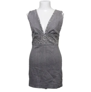 Grå kort klänning med nitar - Snygg grå kortklänning med djup v-ringning och dekorativa nitar längs urringningen och midjan. Klänningen är ärmlös och har en dragkedja i ryggen. Perfekt för dig som vill sticka ut med en edgy look.