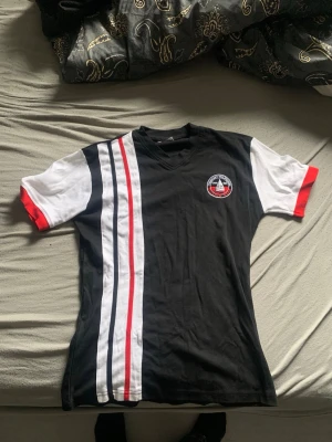 Tommy Hilfiger Sailing Gear t-shirt - Snygg t-shirt från Tommy Hilfiger med Sailing Gear-logga på bröstet. Svart bas med vita och röda detaljer samt ränder längs ena sidan. V-ringad och kortärmad modell, perfekt för en sportig och stilren look.Står Xl men är mer M 