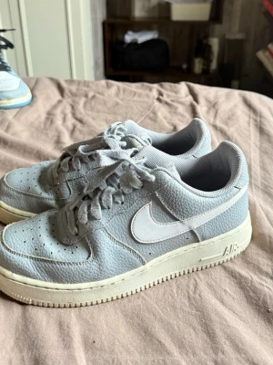Nike Air Force 1 låga sneakers grå - Snygga Nike Air Force 1 sneakers i ljusgrått läder med vit sula och klassisk vit Swoosh på sidan. Skorna har perforerad tå, matchande grå skosnören och en retroinspirerad look. Perfekta för dig som gillar stilrena och tidlösa sneakers.