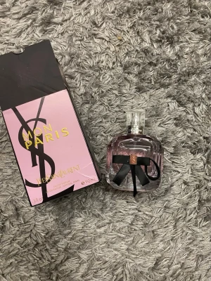 Yves Saint Laurent Mon Paris parfym - Mon Paris från Yves Saint Laurent är en elegant parfym i en lyxig, ljuslila glasflaska med svart rosett och detaljer i roséguld. Flaskan rymmer 90 ml och kommer i en snygg rosa och svart kartong med guldtext. Perfekt för dig som gillar exklusiva dofter och snygg design.