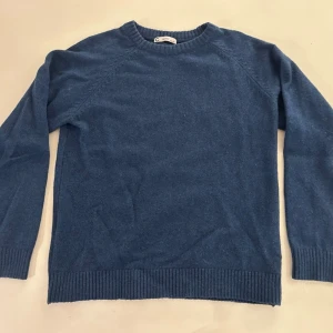 Blå stickad tröja med rund hals - En stilren blå stickad tröja med rund halsringning och långa ärmar. Tröjan har ribbstickade muddar vid ärmslut och nederkant, vilket ger en klassisk och enkel look. Perfekt att matcha med jeans eller kjol.