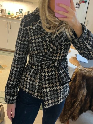 Kort mönstrad jacka - Snygg trenchcoat med klassiskt hundtandsmönster och knytskärp i midjan. Rocken har dubbelknäppning med stora svarta knappar och markerad krage. Passar perfekt till höst och vinter för en trendig look. En knapp är lös.