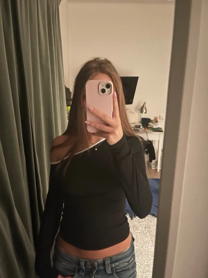 Svart offshoulder topp med vit kant - Cool svart offshoulder topp med lång ärm och vit kant längs halsen. Snygg passform som framhäver axlarna och ger en trendig look. Perfekt att matcha med jeans eller kjol för en stilren outfit.