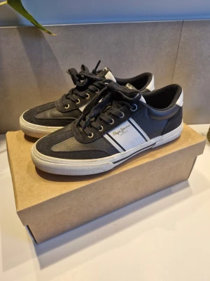 Blåa pepe jeans skor - Snygga svarta sneakers från Pepe Jeans med vita detaljer och logga på sidan. Skorna har klassisk låg profil, svarta skosnören och vit sula. Materialet är en mix av textil och mockaimitation som ger en stilren och sportig look. Boxen tillkommer och om du har frågor eller pruta. Den ser lite smutsig ut på bilden vid dem vita delarna men det är bara att ta bort med vatten eller nåt