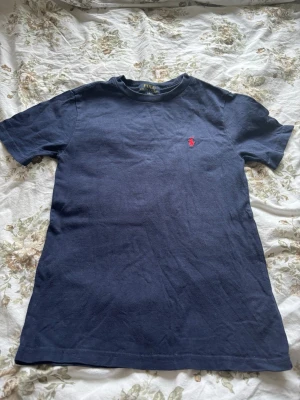 Mörkblå t-shirt från Polo Ralph Lauren - Klassisk mörkblå t-shirt från Polo Ralph Lauren med korta ärmar och rund hals. T-shirten har den ikoniska röda logotypen broderad på bröstet och är gjord i mjuk bomull för en skön känsla. Perfekt till jeans eller shorts för en clean look.