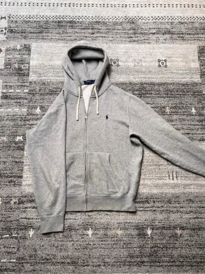 Grå Polo Ralph Lauren hoodie  - Säljer nu denna snygg, stilrena gråa hoodie från Polo Ralph Lauren med dragkedja, huva med vita snören och klassisk broderad logga på bröstet. Mjuk bomullsblandning och känguruficka framtill. Perfekt för en chill och stilren look. Hoodien är självklart äkta och har ej andvänds så mycket! Pris är tyvärr hugget i sten, tveka ej vid frågor och funderingar! Mvh.