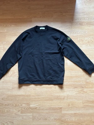 Svart sweatshirt från Stone Island - Svart sweatshirt från Stone Island med klassisk rund hals och lång ärm. Ikonisk logotyp-patch på vänster ärm. Tröjan är i mjukt bomullsmaterial och har ribbade muddar vid ärmslut och nederkant. Perfekt för en stilren och avslappnad look.