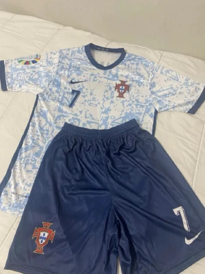 Portugal fotbollskit Nike blå/vit - Snyggt Portugal fotbollskit från Nike med matchtröja i vitt och ljusblått mönster, samt blå shorts. Tröjan har nummer 7 och Portugals landslagsemblem på bröstet. Shortsen är blå med nummer 7 och emblem på benet. Perfekt för fotbollsträning eller match.