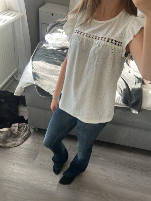 Vit blus med spetsdetalj - Söt vit blus med broderade hål och en dekorativ spetsdetalj över bröstet. Blusen har korta volangärmar och rund halsringning i storlek xs 💞