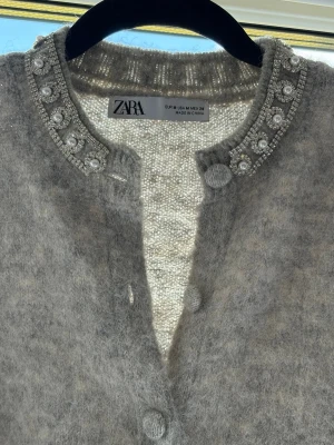 Grå fluffig kofta från Zara med pärlor - Supersnygg grå fluffig kofta från Zara med rund hals och dekorativa pärlor och strassdetaljer längs kragen. Koftan har stora matchande knappar framtill och är gjord i ett mjukt, lurvigt material som känns lyxigt och trendigt.