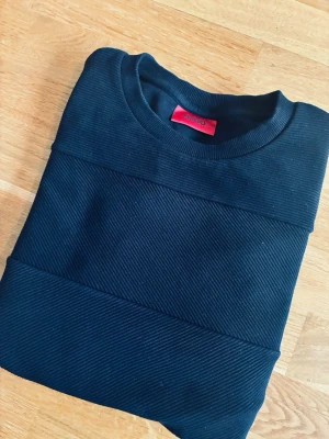 Svart sweatshirt från Hugo - Snygg svart sweatshirt från Hugo med diskreta ribbade paneler på framsidan. Tröjan har rund halsringning och långa ärmar. Perfekt för en stilren och avslappnad look. Materialet känns mjukt och bekvämt mot huden.